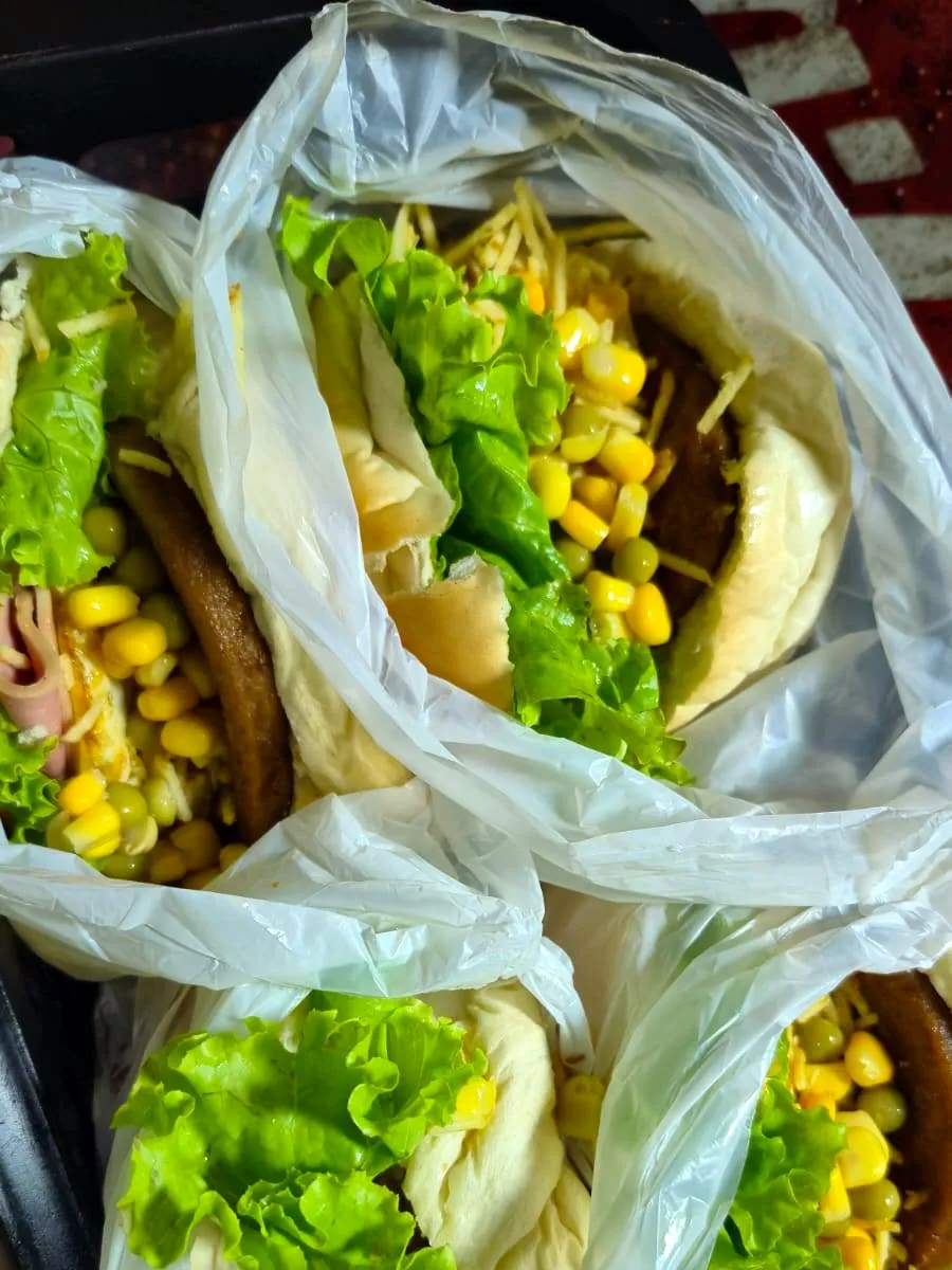 Quatro lanches da Barraca do Baiano vistos de cima, embalados em sacos plásticos, com alface, milho, ervilha e batata palha à mostra